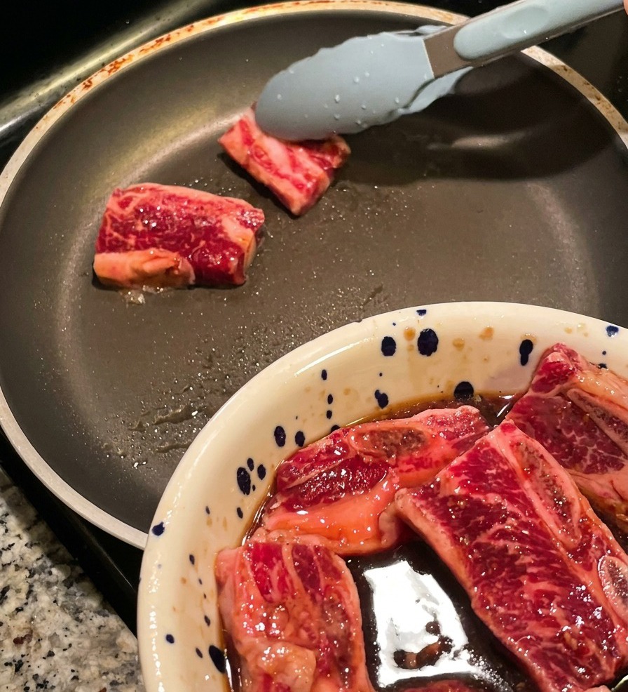 Unas pinzas colocando tiras de costillas de res marinadas en una sartén caliente.