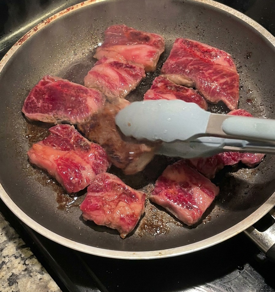 Unas pinzas dando la vuelta a las costillas doradas en una sartén, mostrando una costra oscura y caramelizada.
