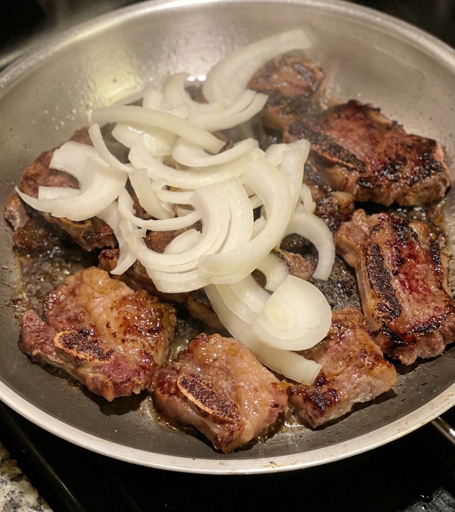 Un puñado de cebolla blanca cruda en juliana sobre unas costillas de res doradas en una sartén de acero inoxidable.