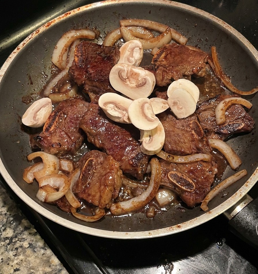 Champiñones en rodajas salteándose con costillas de res y cebolla caramelizada en una sartén oscura.