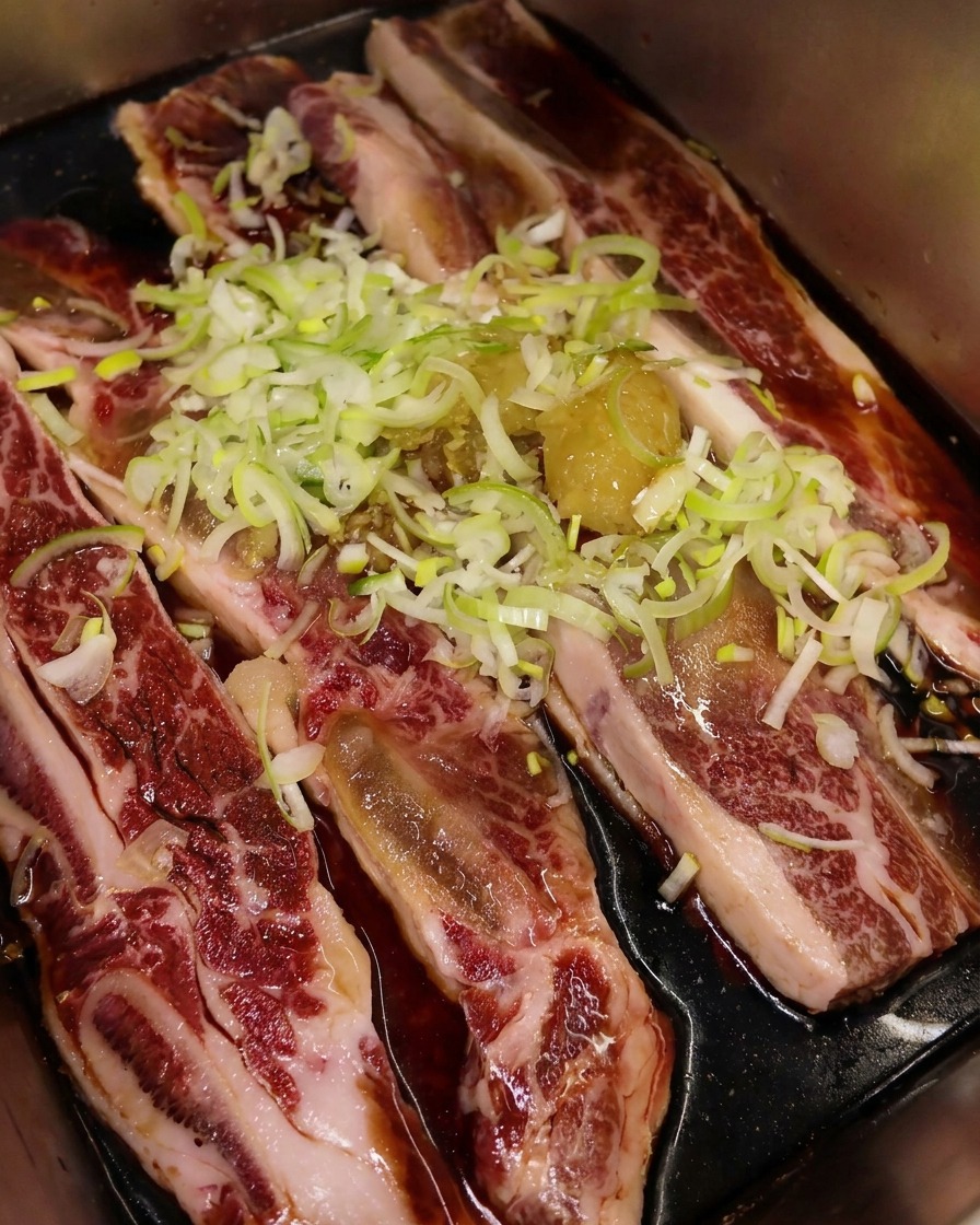 Aceite de sésamo tostado vertiéndose sobre costillas con cebolleta y ajo, creando una capa brillante.