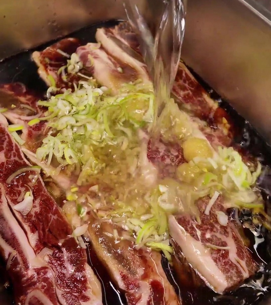 Chorro de agua vertiéndose en una bandeja de acero inoxidable con costillas, ajo y cebolleta.