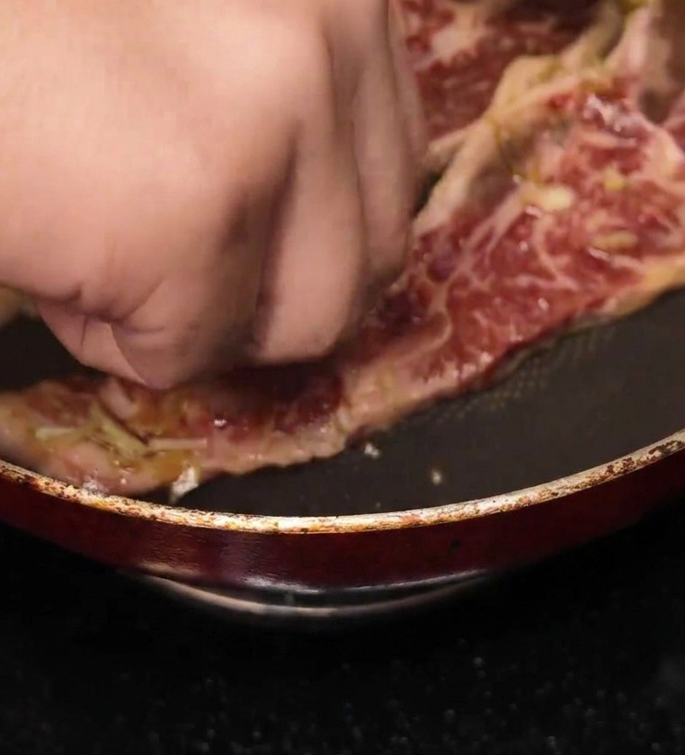 Una mano colocando una tira de costilla marinada en una sartén caliente antiadherente.