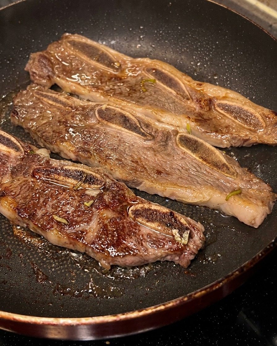 Tres tiras de costillas LA chisporroteando en una sartén negra, una de ellas volteada mostrando la costra oscura.