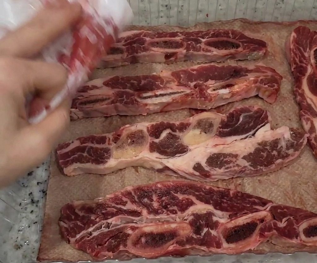 Una mano usando una servilleta de papel blanca para secar tiras de costilla de ternera crudas en una bandeja.
