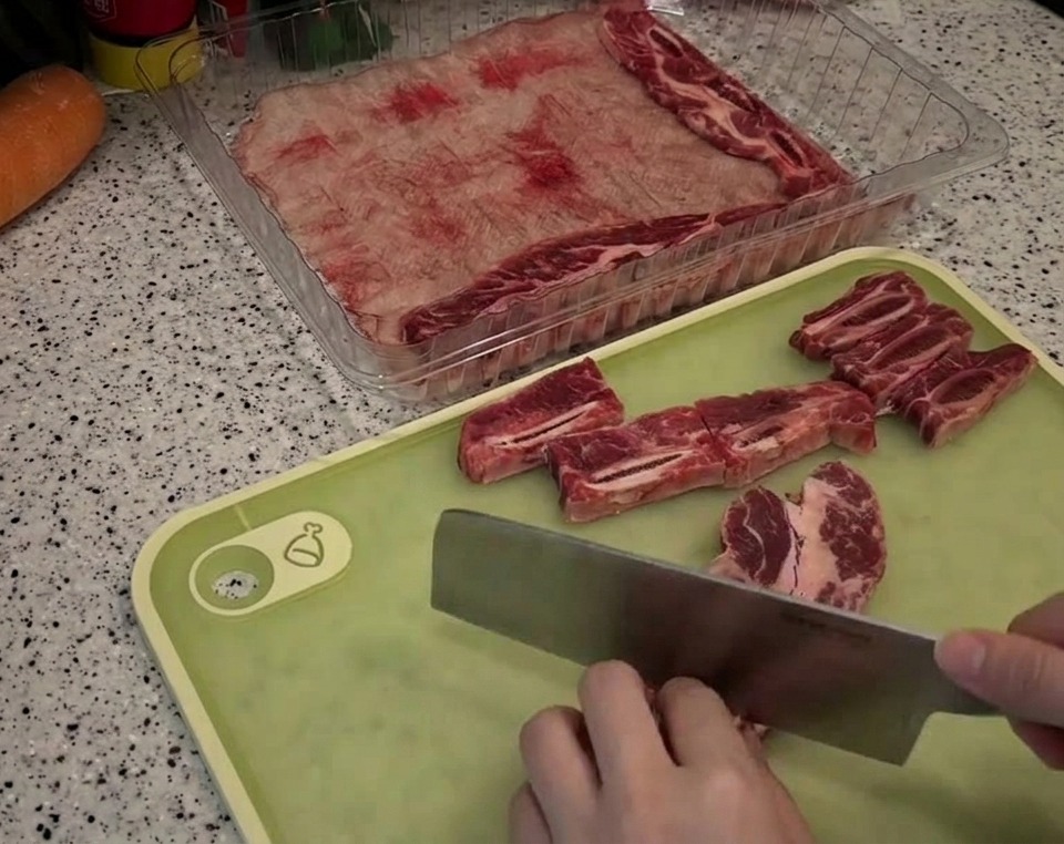 Una persona usando un cuchillo grande para cortar costillas de ternera con grasa veteada en trozos rectangulares sobre una tabla verde.