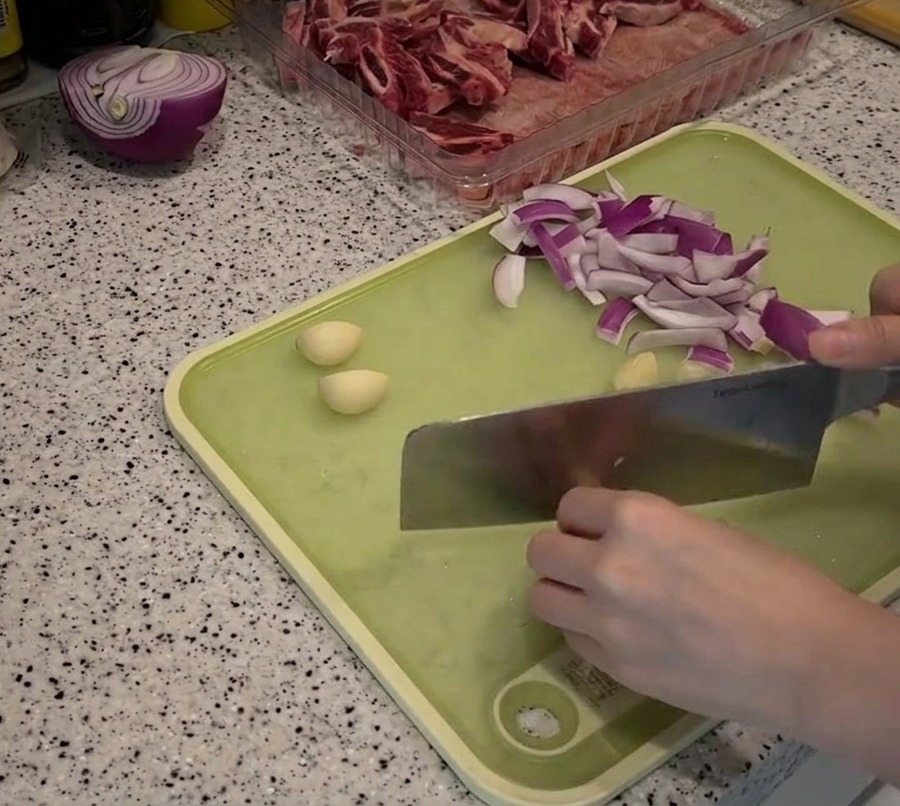 Tiras de cebolla roja y dientes de ajo pelados extendidos sobre una tabla de cortar verde siendo preparados con un cuchillo.