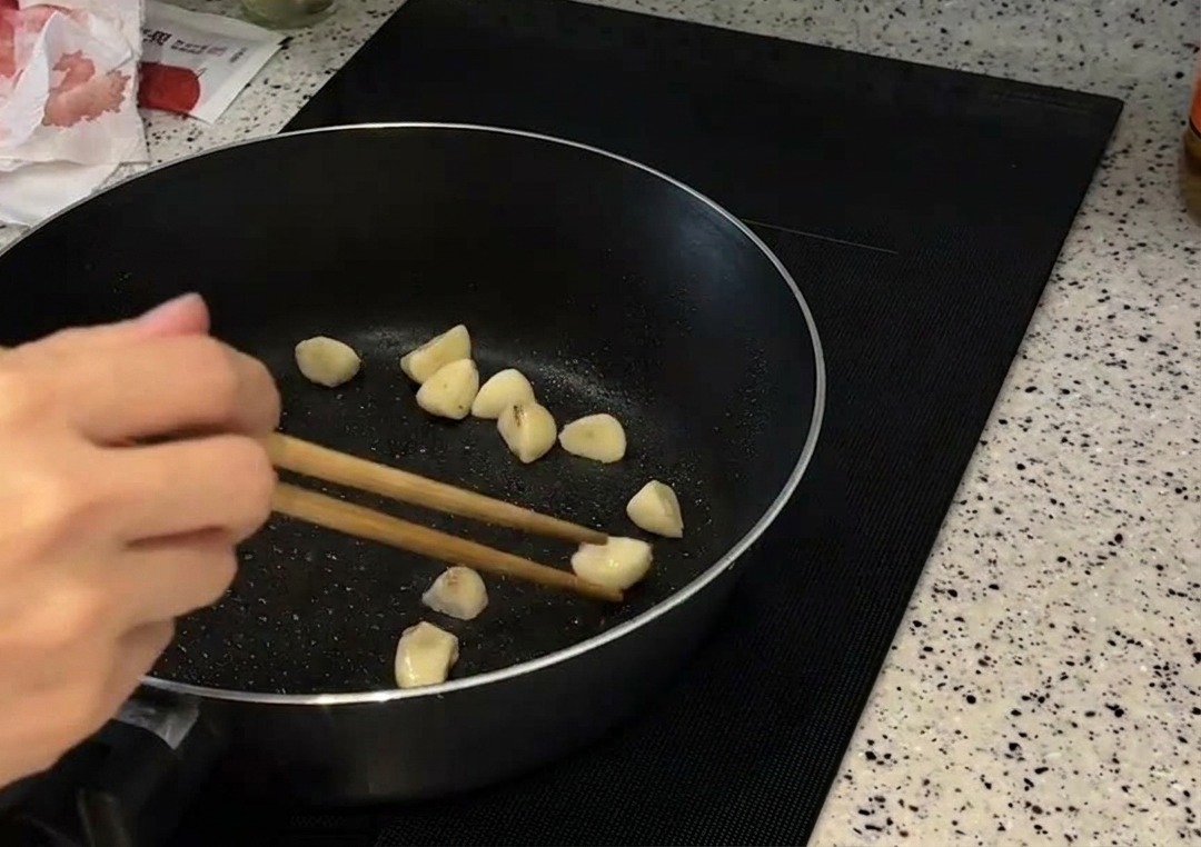 Dientes de ajo enteros siendo removidos con palillos de madera en una sartén oscura hasta que se vuelven dorados.