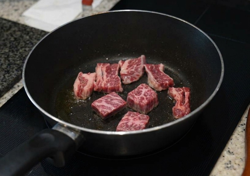 Varios trozos de costilla de ternera veteada sellándose en una sartén oscura, con los bordes tornándose marrones.