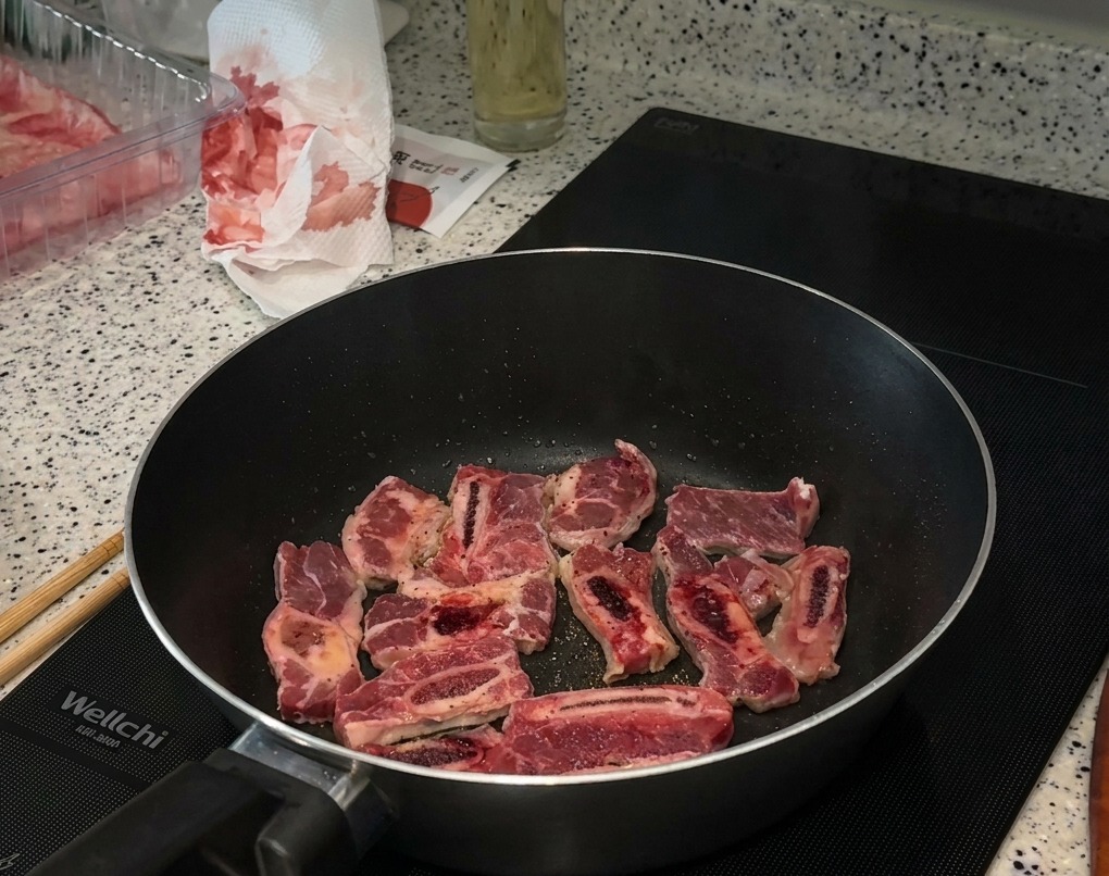 Trozos de costilla de ternera sellándose en una sartén sobre una placa de inducción, con sal y pimienta visible sobre la carne.