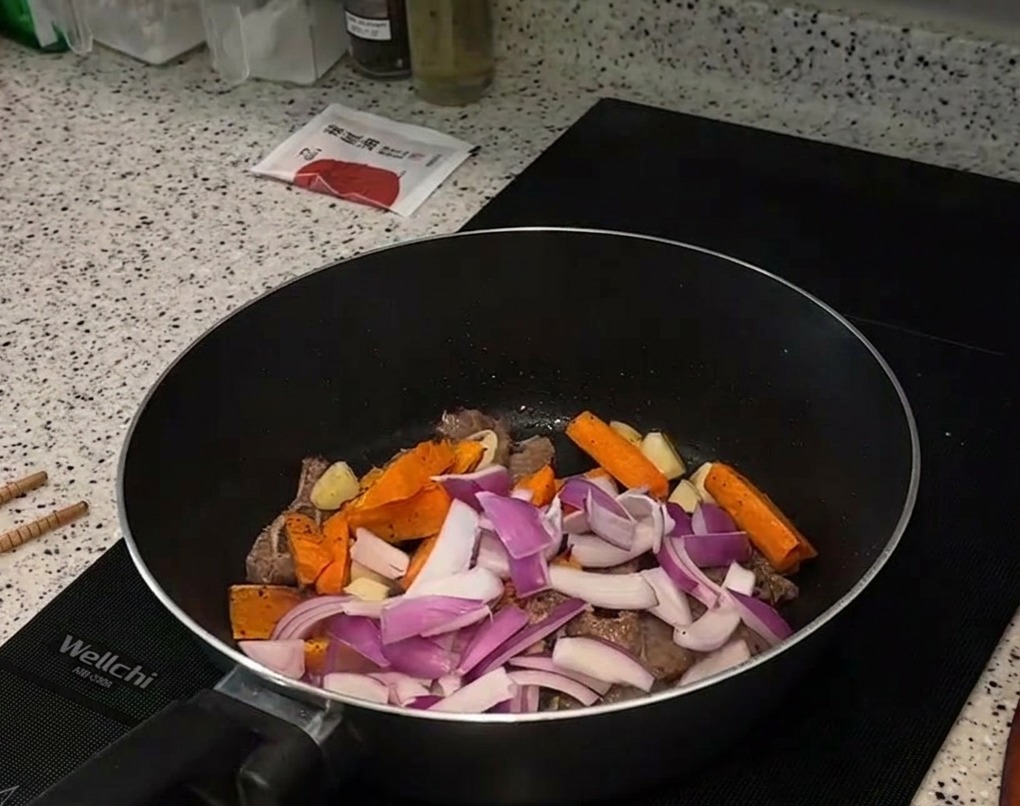 Tiras de cebolla roja y bastones de zanahoria asada siendo añadidos sobre los trozos de ternera sellados en una sartén.