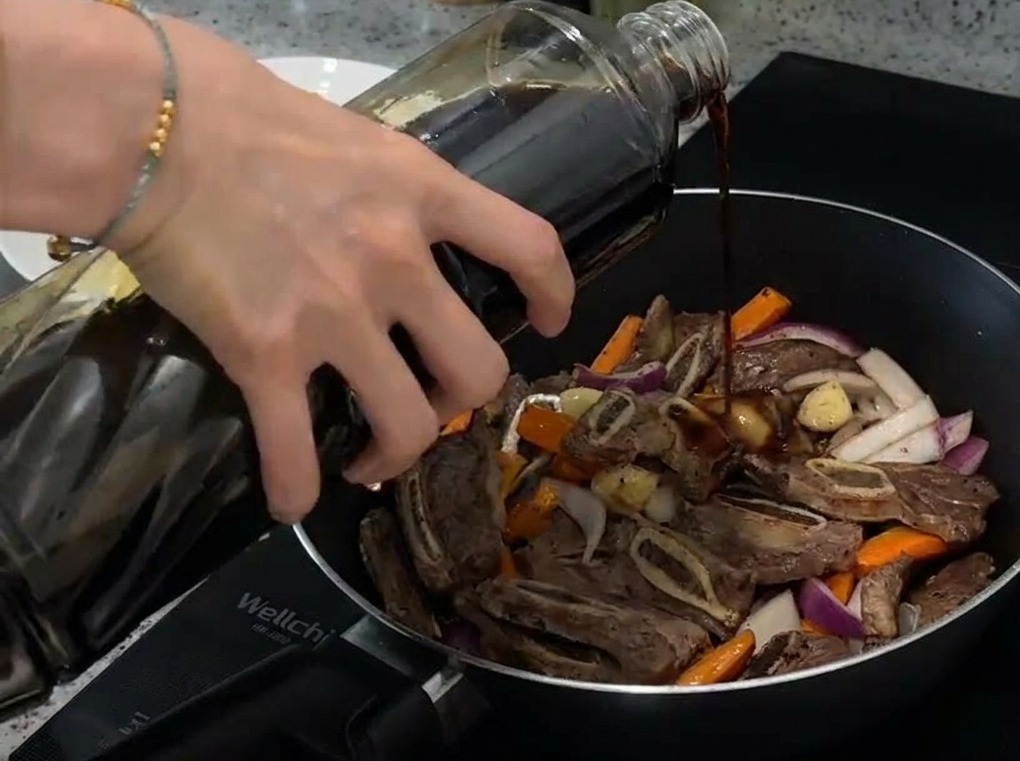 Primer plano de salsa de soja clara siendo vertida desde una botella de vidrio a una sartén con carne, zanahorias y cebolla.
