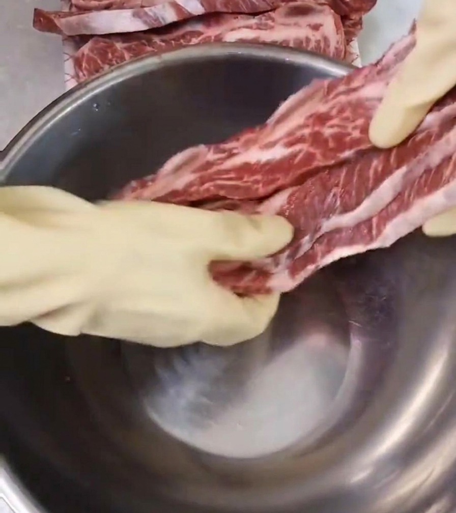 Una persona con guantes blancos colocando costillas de res crudas y marmoleadas en un bol hondo de acero inoxidable sobre una encimera limpia.