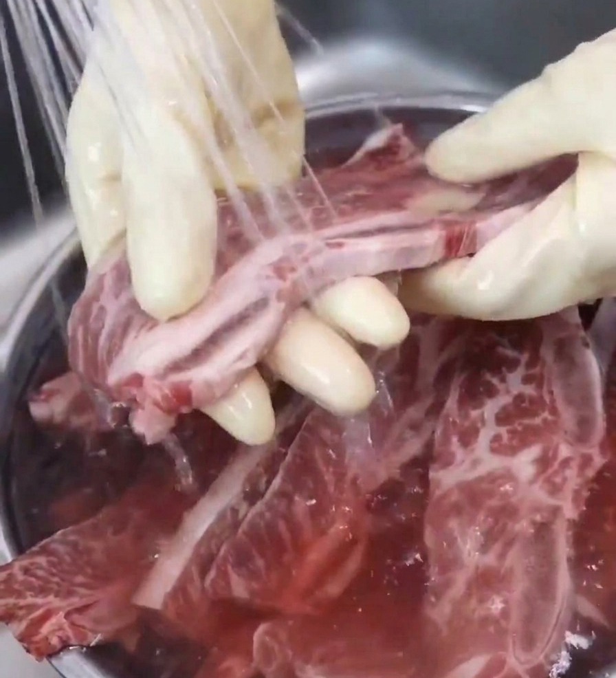 Una mano con guante sostiene una costilla de res bajo el chorro de agua del grifo, enjuagándola sobre un bol para eliminar las impurezas.