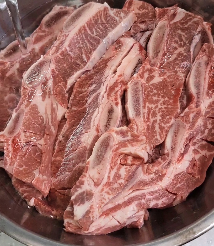 Agua limpia cayendo en un bol de acero inoxidable hasta cubrir una gran cantidad de costillas de res crudas previamente marcadas.