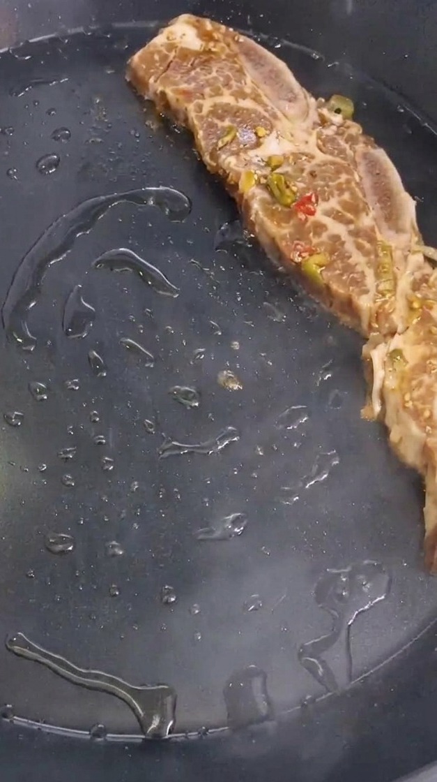 Una costilla de res marinada, sazonada con trocitos de pimiento y cebolla, colocándose sobre una sartén negra caliente y engrasada.