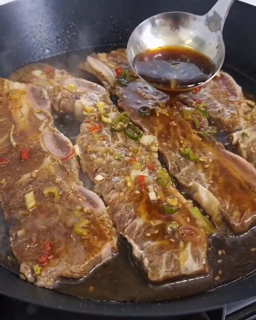 Un cazo de acero inoxidable vertiendo una marinada oscura sobre varios trozos de costillas de res que se cocinan en una sartén grande.