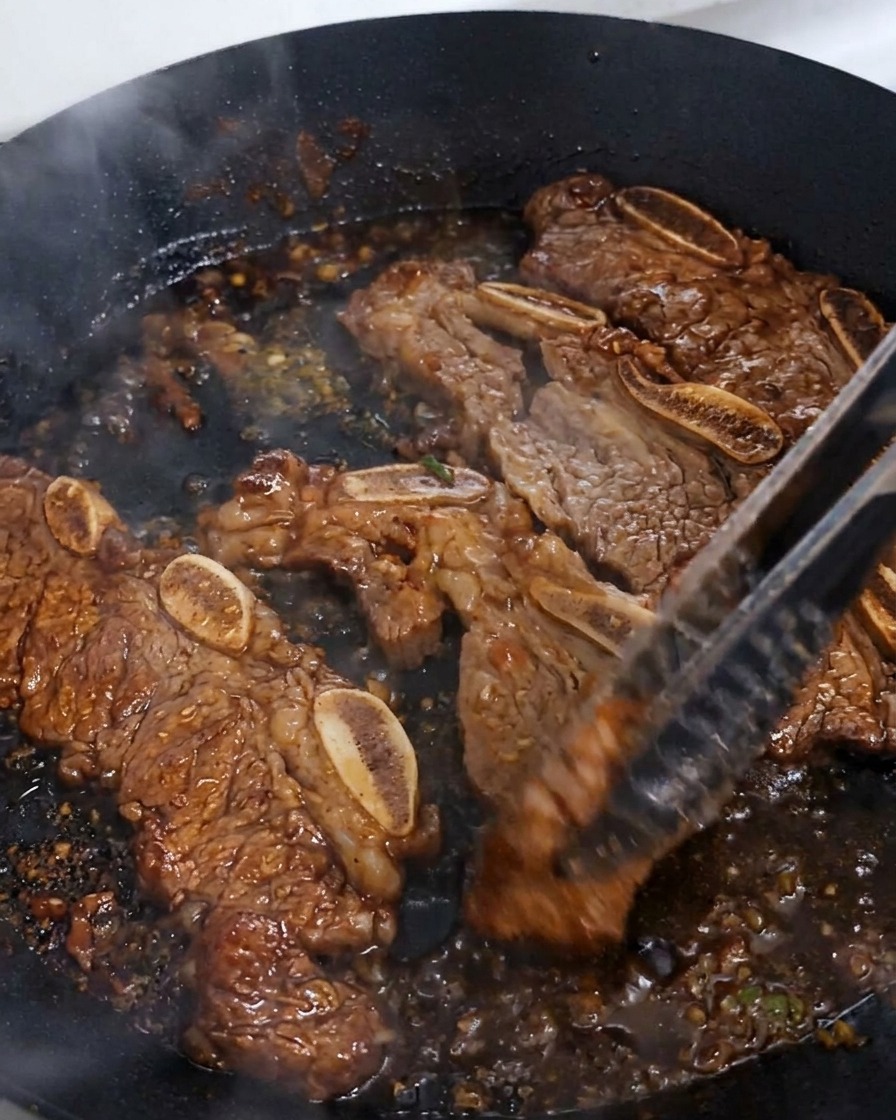 Costillas de res siendo volteadas con unas pinzas en una sartén donde la salsa oscura está burbujeando y reduciéndose para formar un glaseado espeso y brillante.