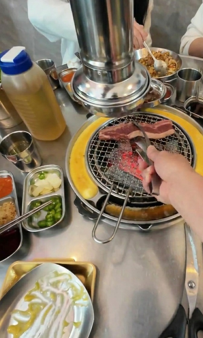 Unas pinzas metálicas colocando tiras de carne de ternera sobre una parrilla de carbón caliente rodeada de guarniciones.