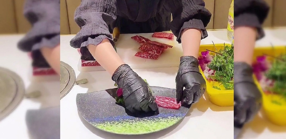 Manos con guantes negros colocando cuidadosamente láminas de ternera veteada en forma de abanico superpuesto sobre un plato oscuro curvo.