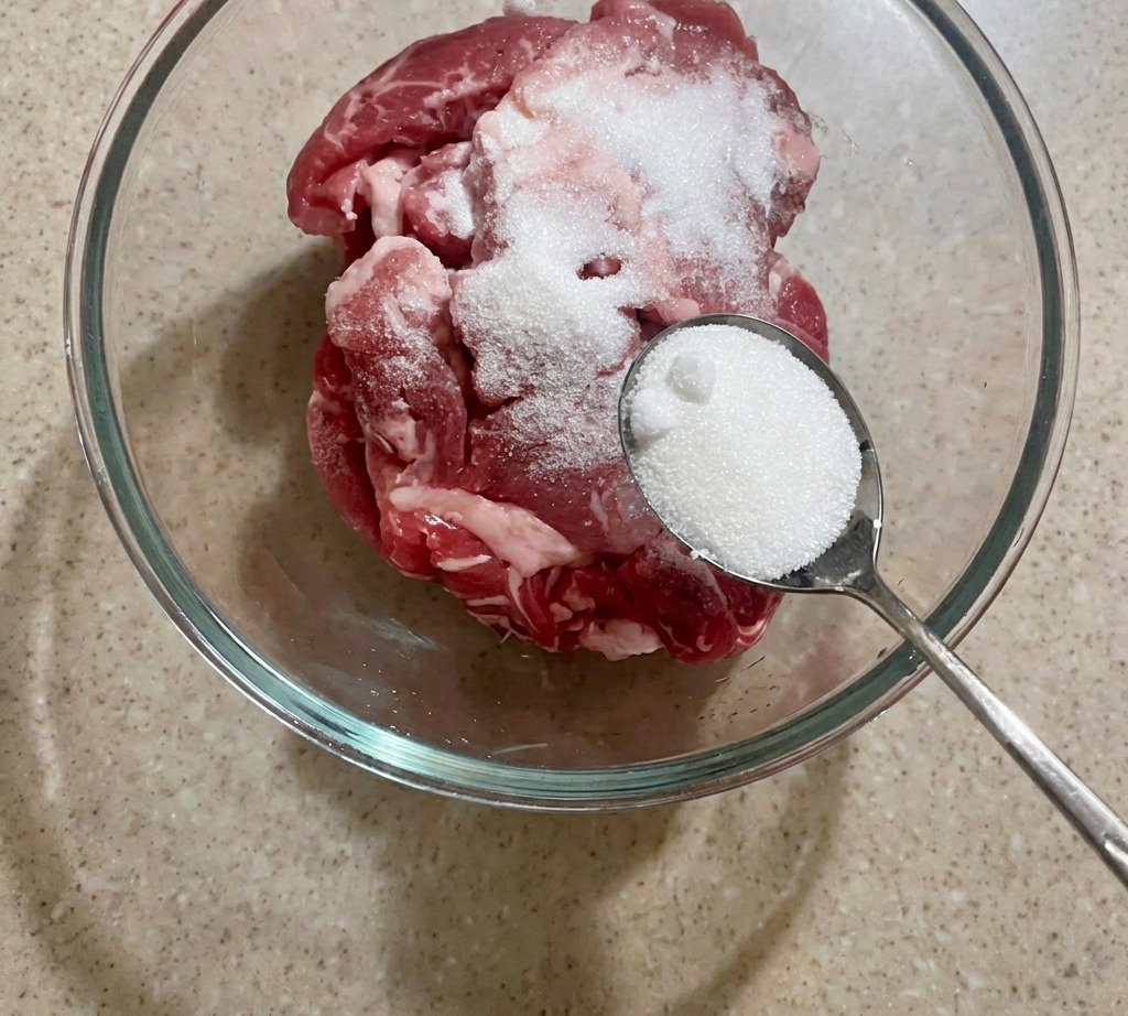 Un bol con carne de cerdo en láminas siendo sazonada con una cucharada de azúcar blanca.