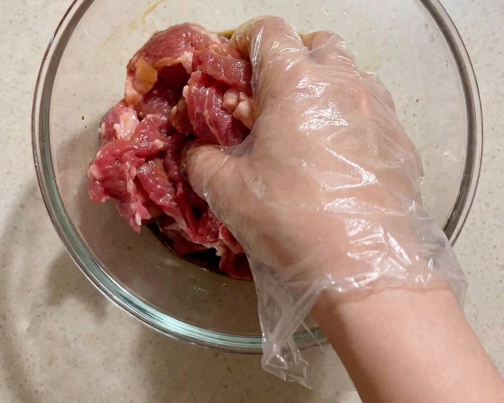 Una mano con guante transparente mezclando láminas de cerdo crudo con condimentos en un bol de cristal.