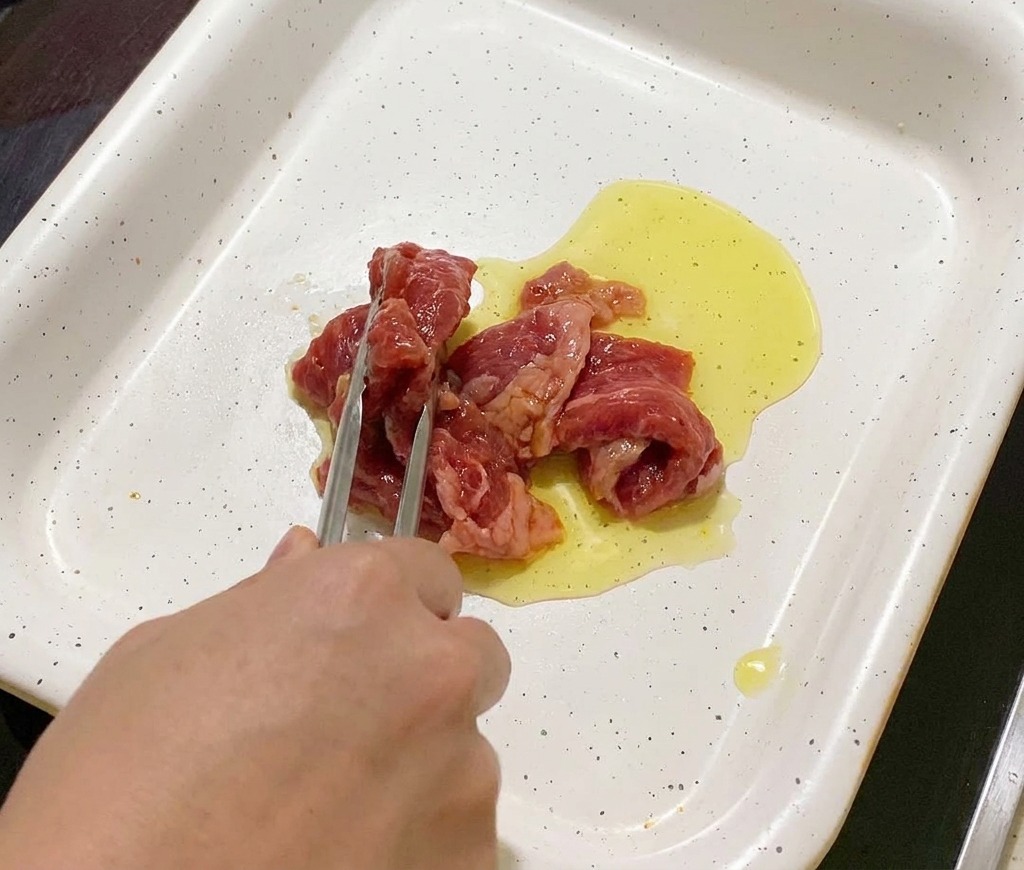 Unas pinzas colocando láminas de cerdo marinado en una sartén blanca con aceite caliente.