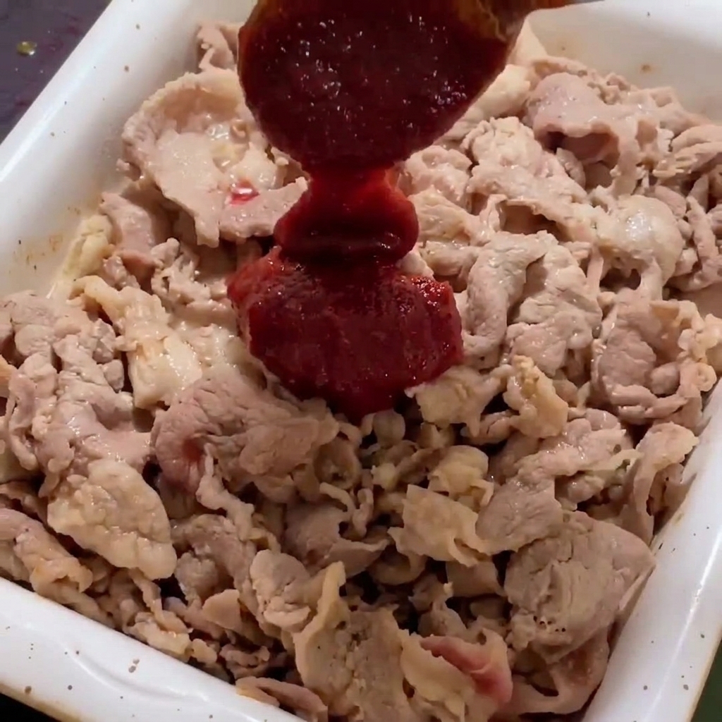 Salsa roja picante y espesa añadiéndose a las láminas de cerdo doradas en una sartén blanca.
