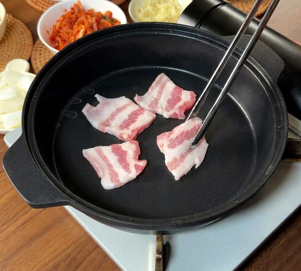 Tiras finas de panceta de cerdo cruda y veteada colocándose en una sartén negra con palillos largos de metal.