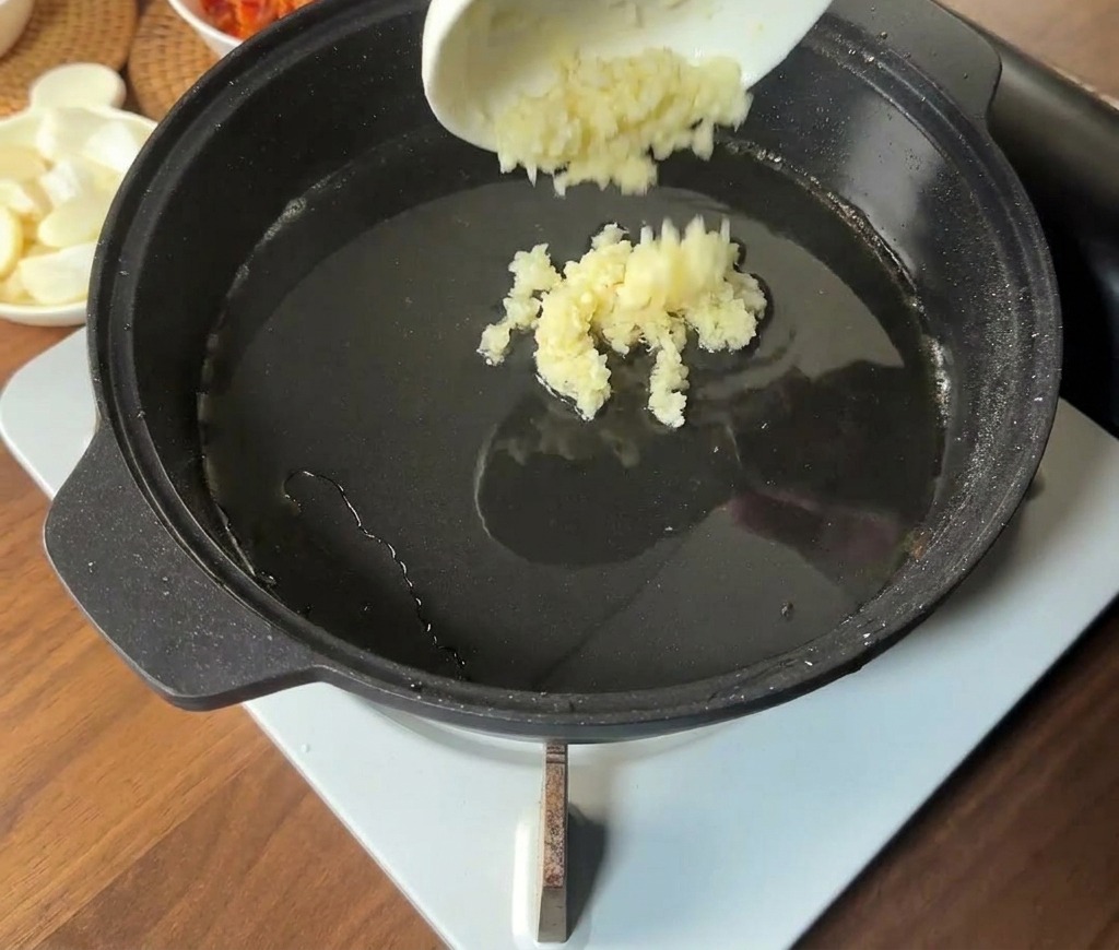 Ajo picado vertiéndose desde un pequeño cuenco blanco al centro caliente y aceitoso de una sartén negra.