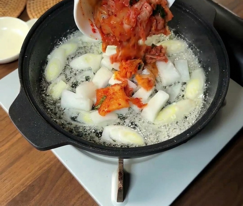 Kimchi coreano picado y rodajas de cebolla blanca añadiéndose a una sartén con cebolletas sofritas.