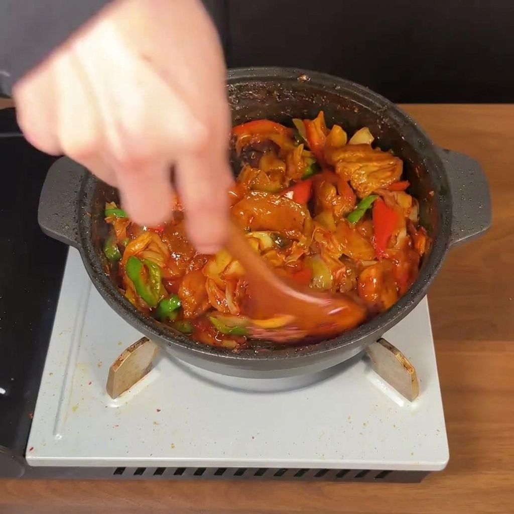 Una cuchara de madera removiendo vigorosamente una sartén con panceta, kimchi y chiles en una espesa salsa roja.