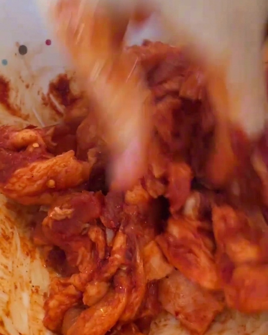 Una mano con guante masajeando la carne con la salsa roja vibrante en un bol blanco.