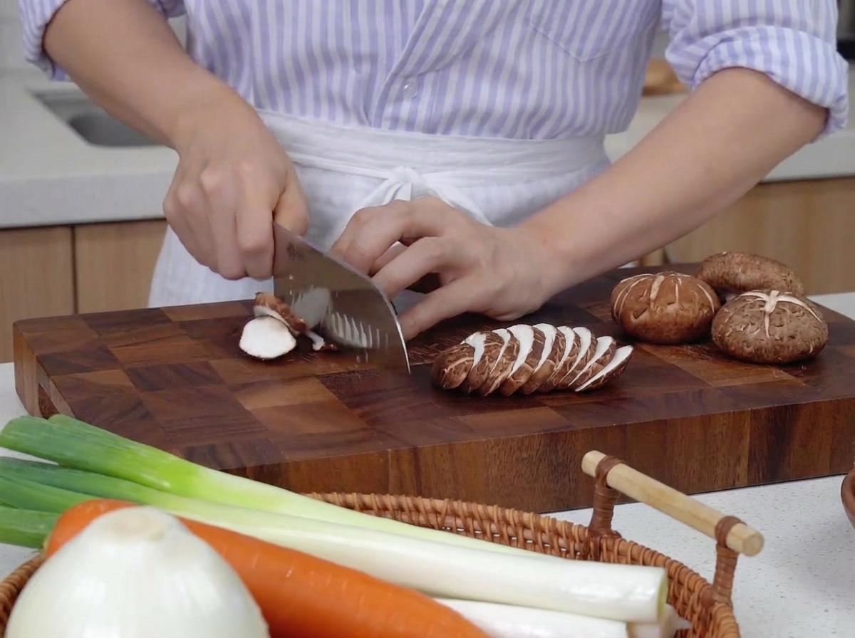 Manos usando un cuchillo de chef para cortar setas shiitake en láminas finas sobre una tabla de madera oscura junto a zanahorias y cebolletas.