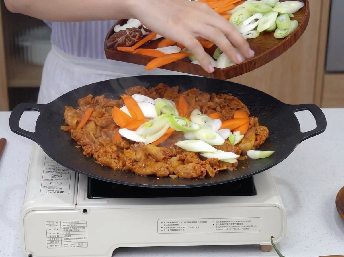 Variedad de verduras laminadas, incluyendo cebolla, zanahoria y setas, cayendo sobre el cerdo picante chisporroteante en una sartén grande.