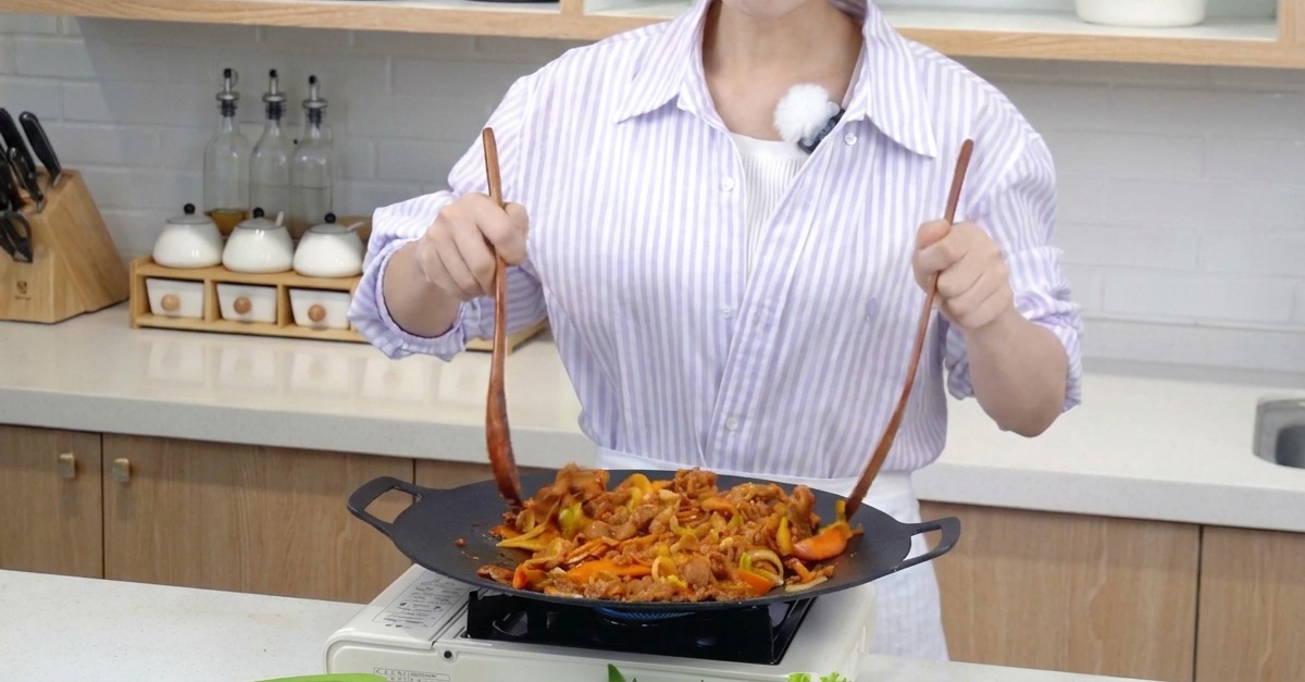 Tiras de cerdo marinado y vegetales coloridos siendo removidos en una sartén amplia con dos espátulas largas de madera.
