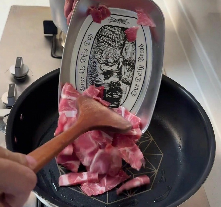 Láminas finas de paleta de cerdo cruda pasando de una bandeja metálica a una sartén caliente con una espátula de madera.