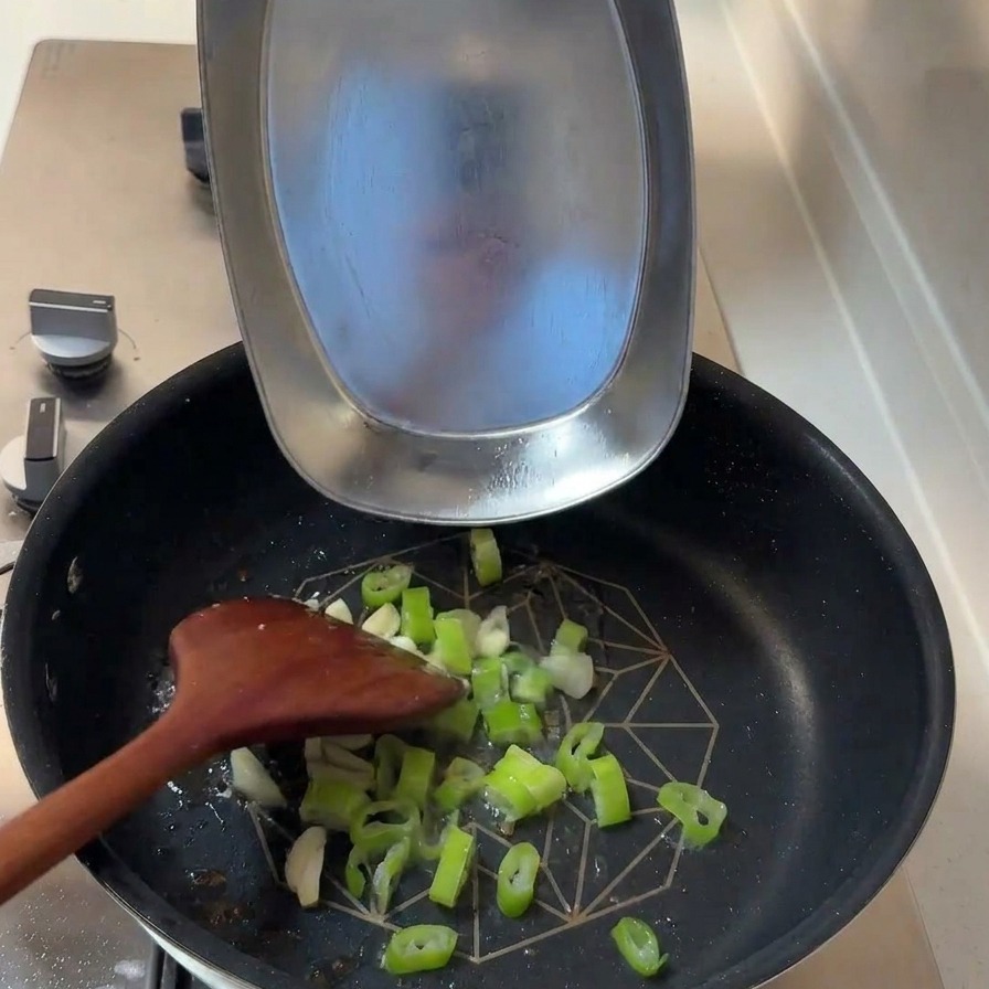 Cebolleta picada y dientes de ajo laminados añadiéndose a una sartén con restos de aceite.
