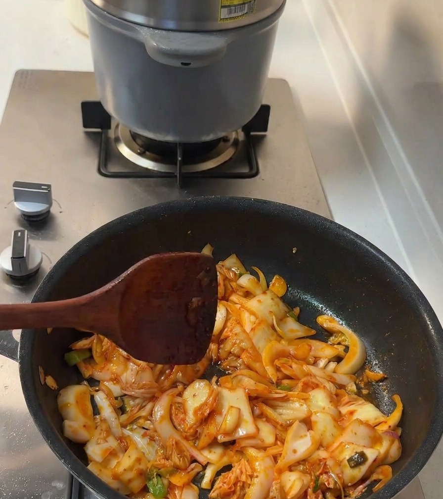 Sazón líquida y azúcar granulada mezclándose con el kimchi y las cebollas en la sartén.