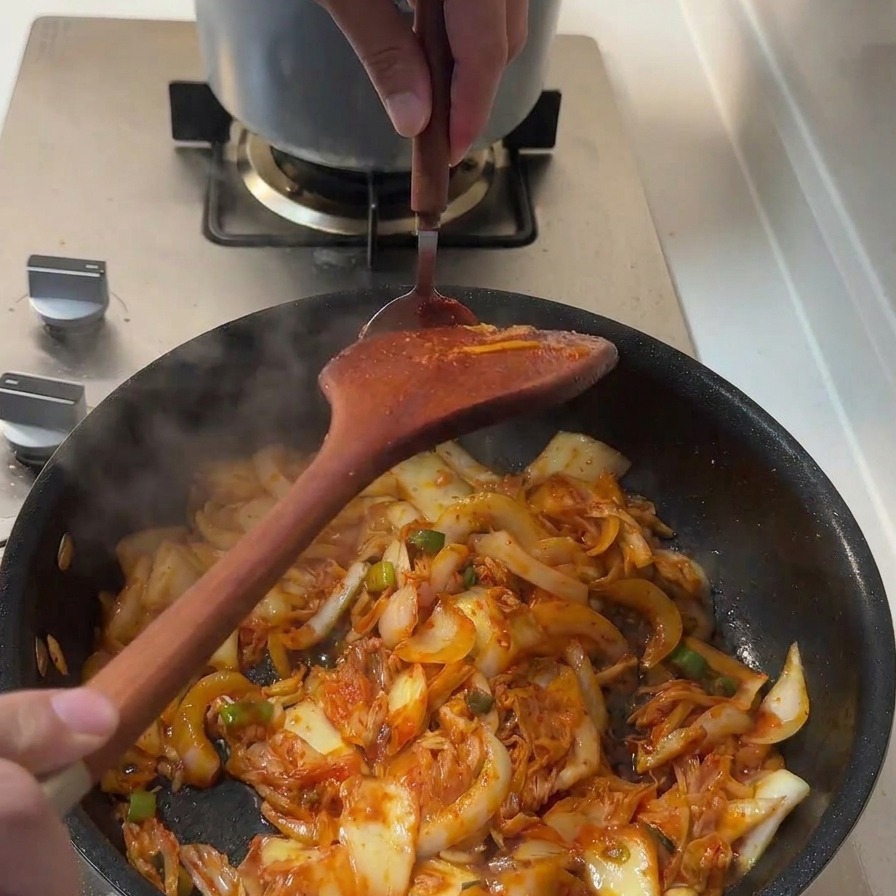 Una cucharada de pasta espesa roja gochujang siendo incorporada a los ingredientes en la sartén.