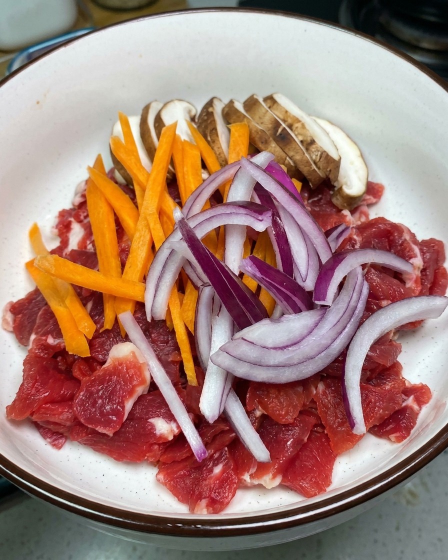 Un bol blanco con láminas de ternera cruda, setas shiitake, zanahoria y cebolla roja.