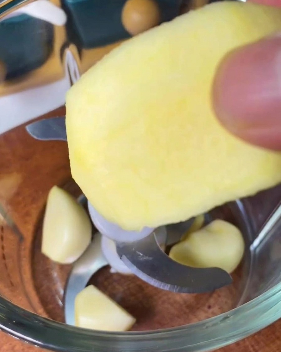 Una persona añadiendo una rodaja de manzana pelada a una batidora con varios dientes de ajo.