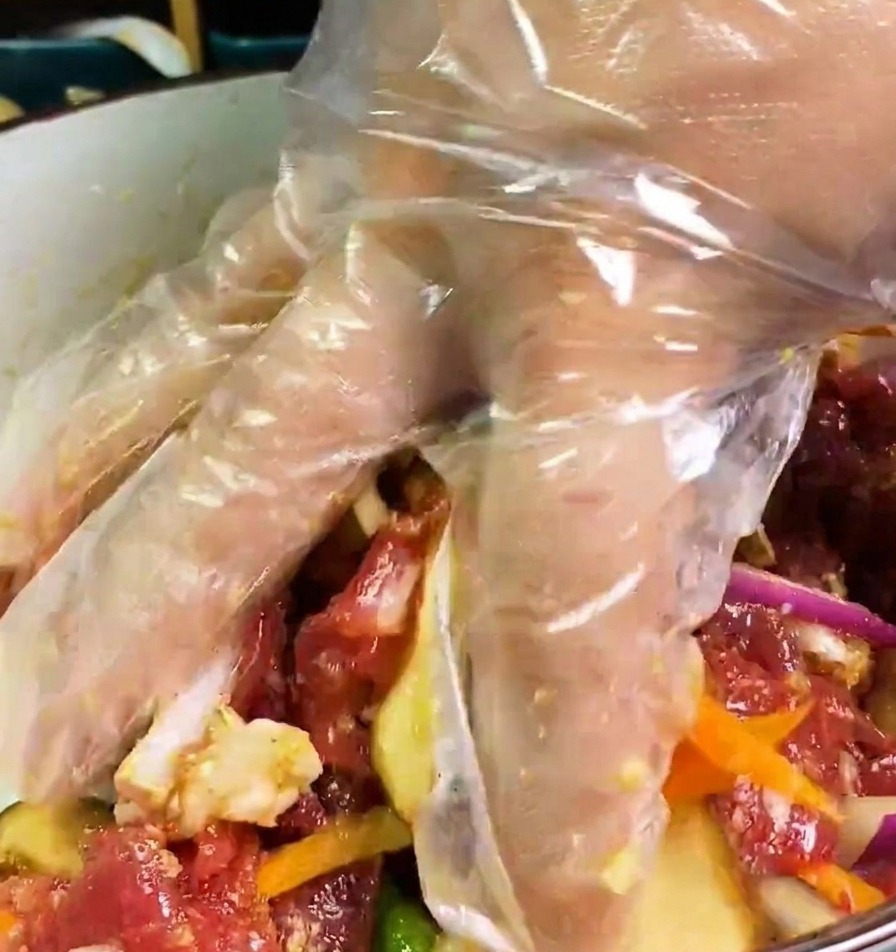 Una mano con guante transparente mezclando ternera, cebolla, zanahoria y setas en un marinado oscuro.