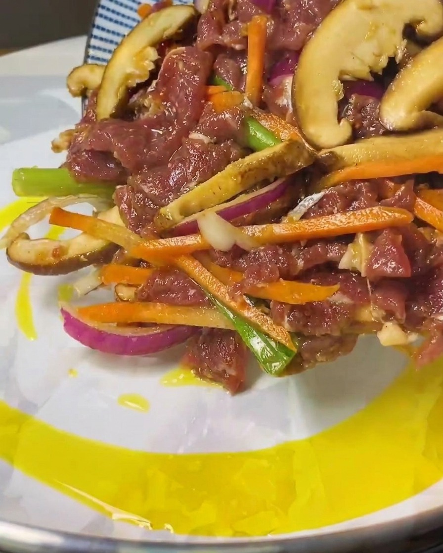 Carne marinada y tiras de verduras deslizándose desde un plato azul a una sartén con aceite caliente.