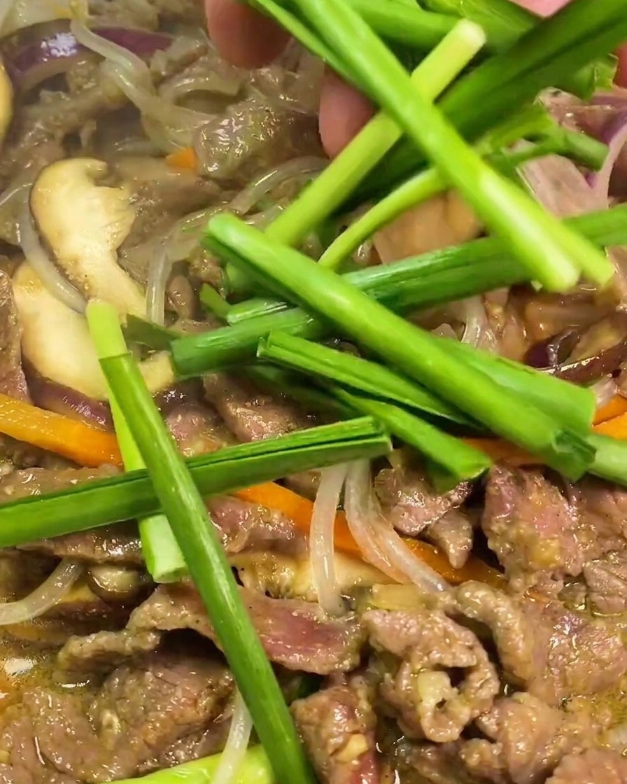 Cebollino chino verde fresco añadiéndose a una sartén con carne, fideos translúcidos, zanahoria y cebolla.
