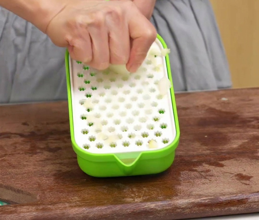 Una persona utilizando un rallador verde para convertir una manzana pelada en pulpa sobre una tabla de madera.