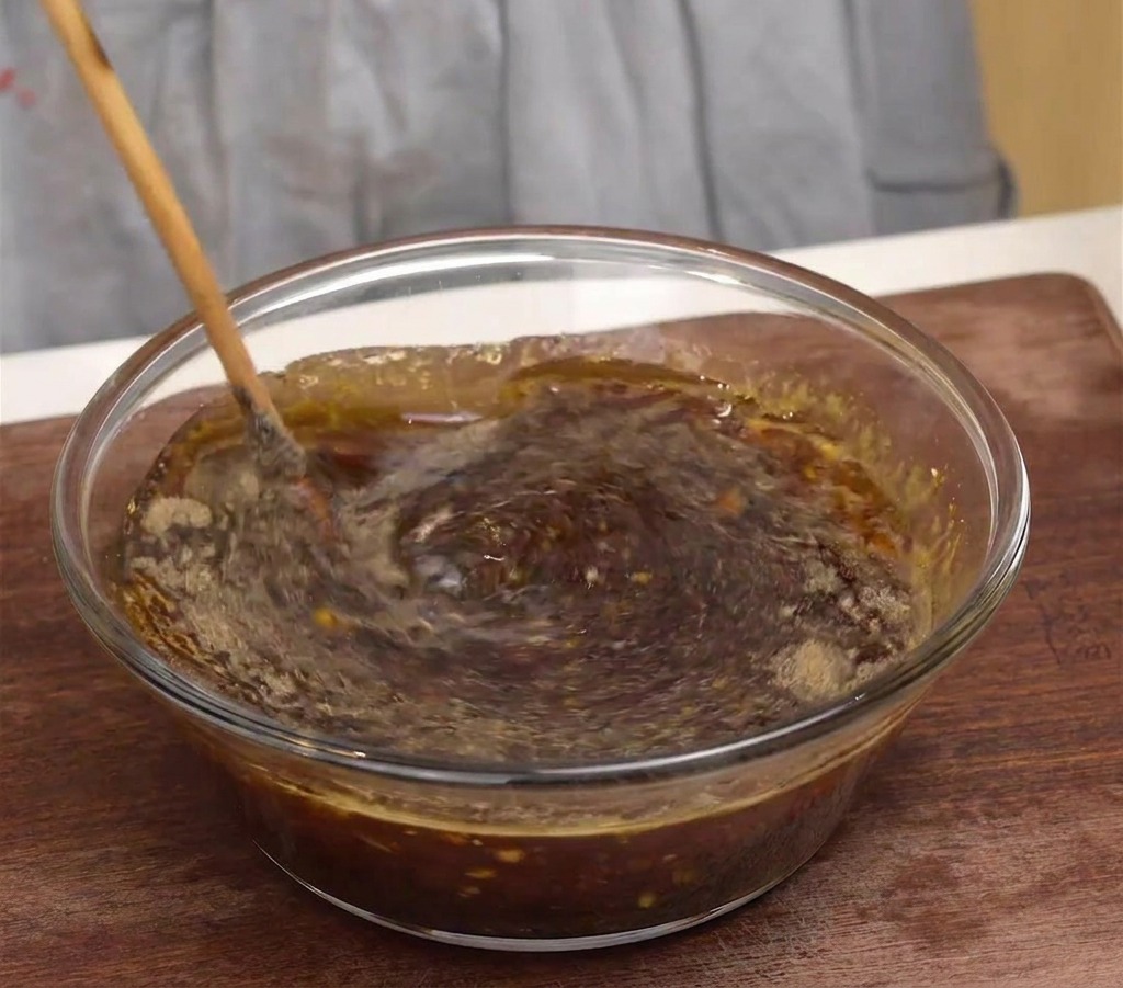 Una cuchara de madera removiendo un marinado oscuro y aromático en un bol de cristal.