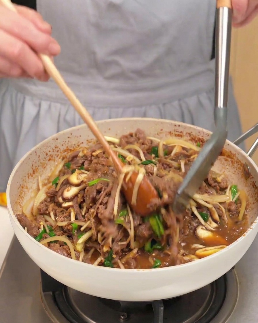 Una sartén con ternera cocinada, setas, cebollas y cebolletas mezclándose con una cuchara de madera.