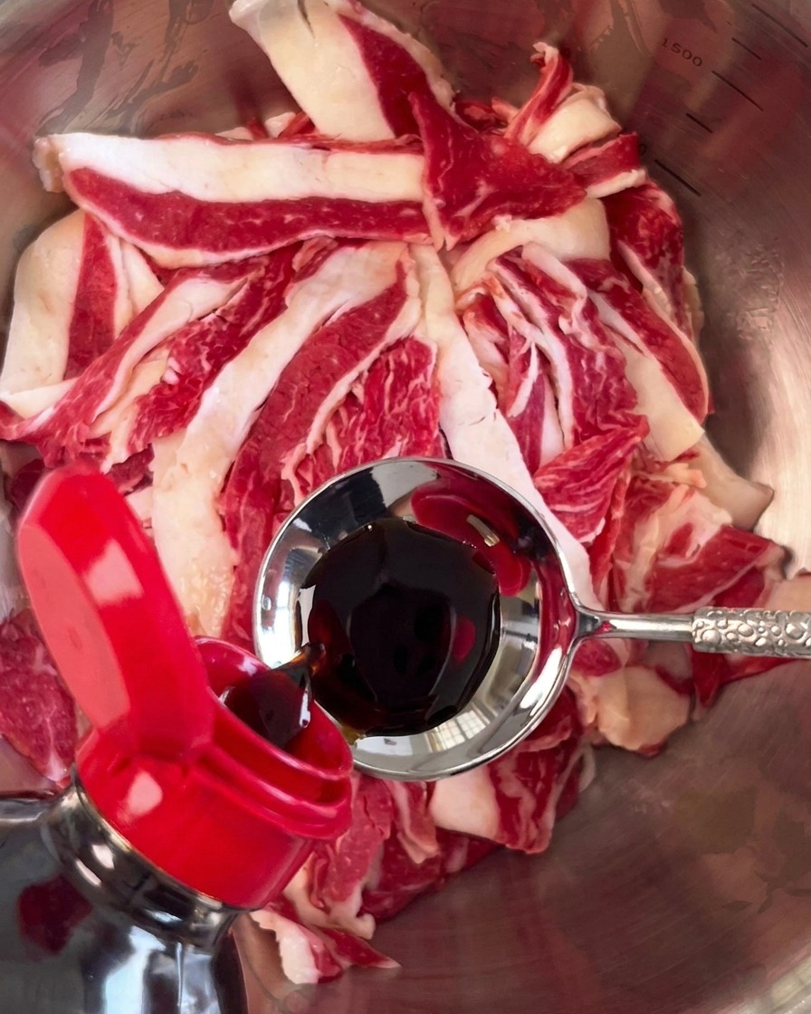 Salsa de soja oscura vertiéndose desde una cuchara sobre la carne cruda en un bol plateado.