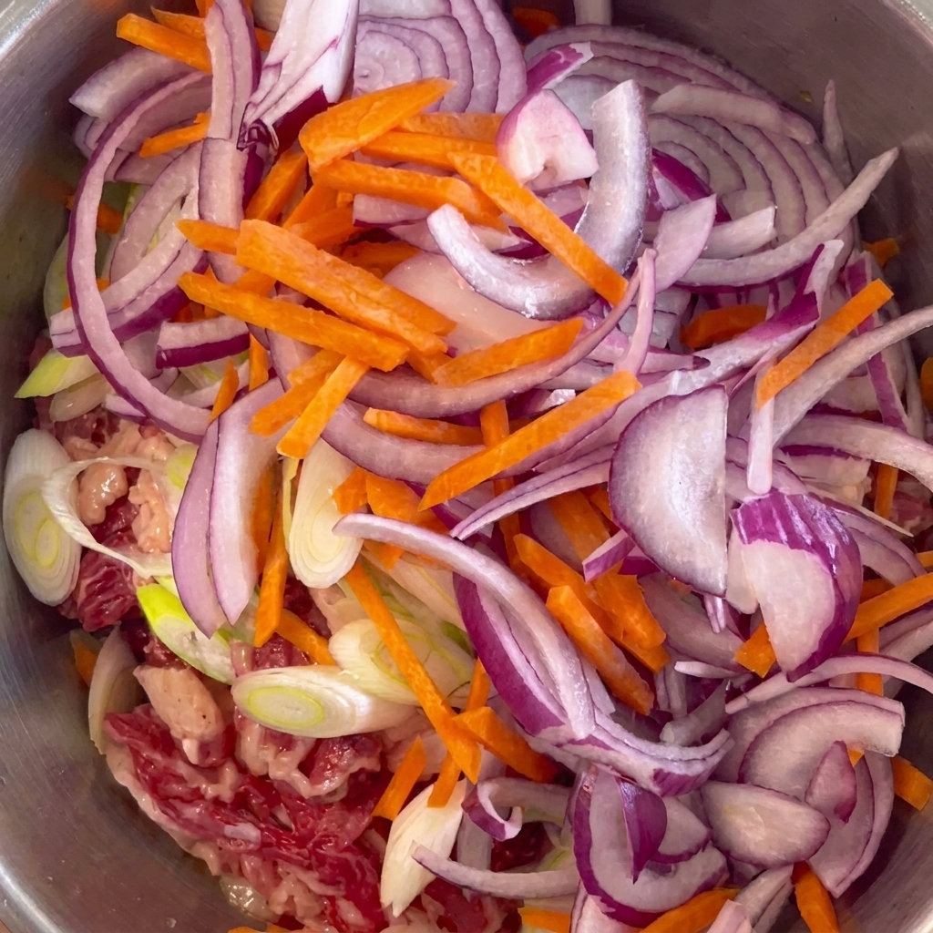 Cebolla morada laminada y bastoncillos de zanahoria sobre la carne marinada en un bol de acero.